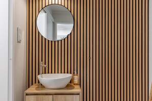 Une salle de bain avec un mur recouvert de lambris en bois, un miroir rond, un lavabo blanc et un distributeur de savon.