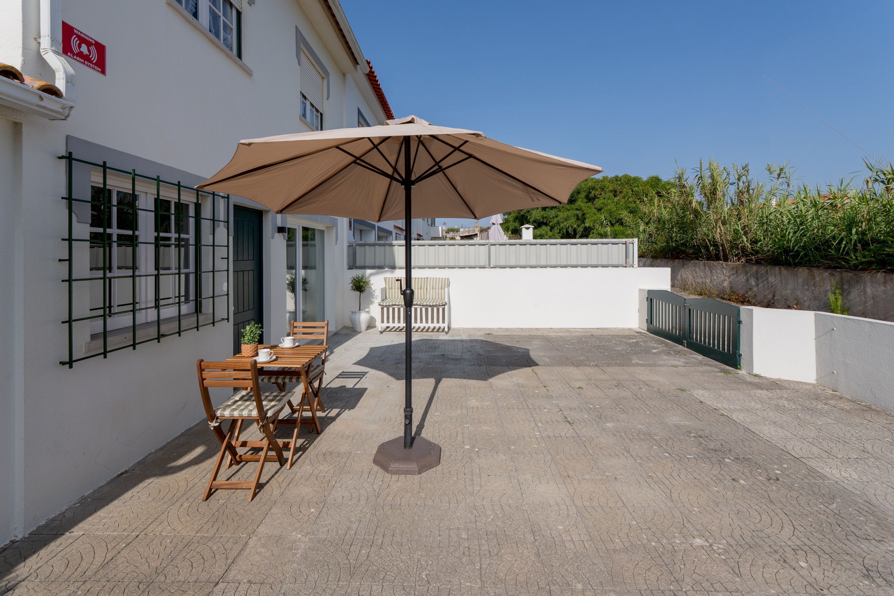 An outdoor patio with a table and chairs, a large umbrella, and a white building with a metal fence.