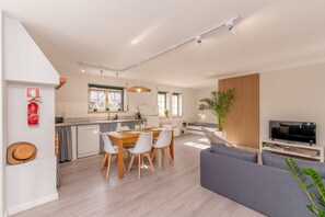 Une cuisine moderne avec des meubles blancs, un coin repas avec une table en bois et des chaises blanches, un canapé gris et un téléviseur posé sur un meuble blanc.