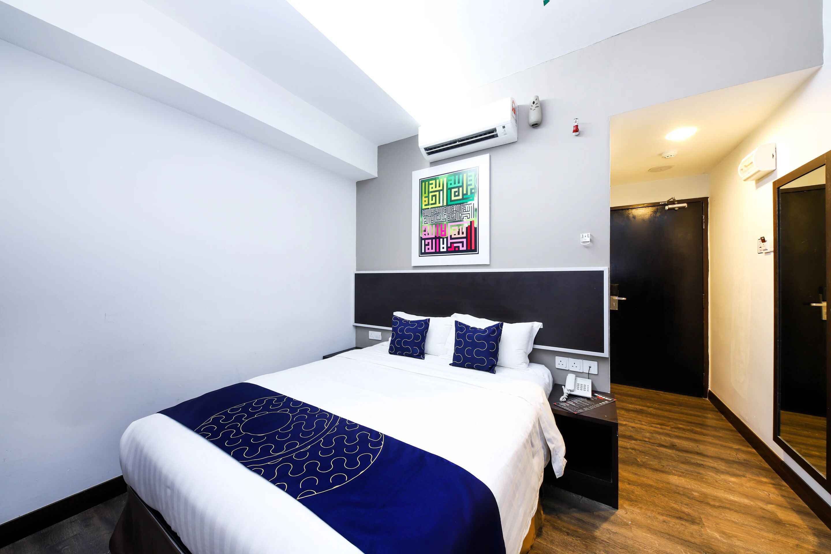 A hotel room with a bed, a desk, an air conditioning unit, a wall-mounted artwork, and a door.