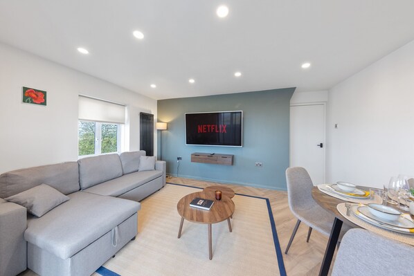 Ett modernt vardagsrum med en grå soffa, ett soffbord och en matplats med ett bord dukat för fyra personer. En tv som är monterad på väggen visar "NETFLIX".