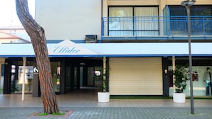 A building with a storefront named 'Ellidor,' featuring a closed shutter, potted plants, and a tree in the foreground. - Apartment 'Ellidor 01' with Private Terrace and Air Conditioning (Lido di Jesolo)