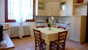 A kitchen with wooden cabinets, a dining table with chairs, a stove, and a window with curtains. - Apartment 'La Pace 02' with Air Conditioning (Lido di Jesolo)