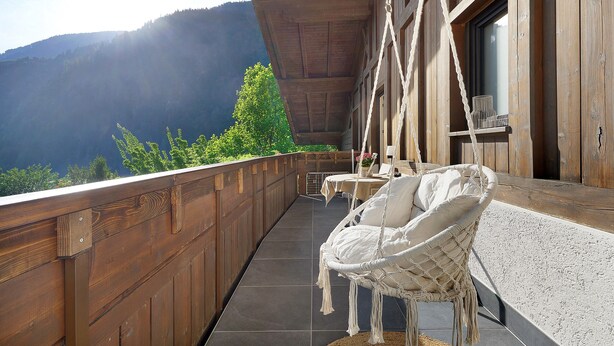 Un balcon en bois avec un fauteuil suspendu, une table et une vue sur les montagnes.