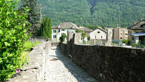 A narrow stone pathway leading to traditional buildings with stone walls and a mountainous backdrop.