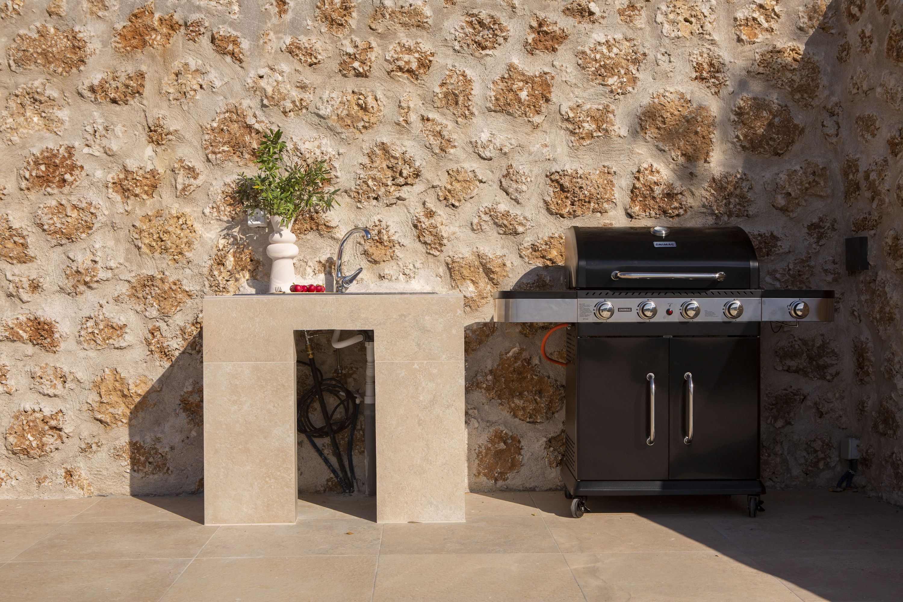 Una parrilla con un muro de piedra de fondo y una pequeña mesa con una planta y un objeto rojo.