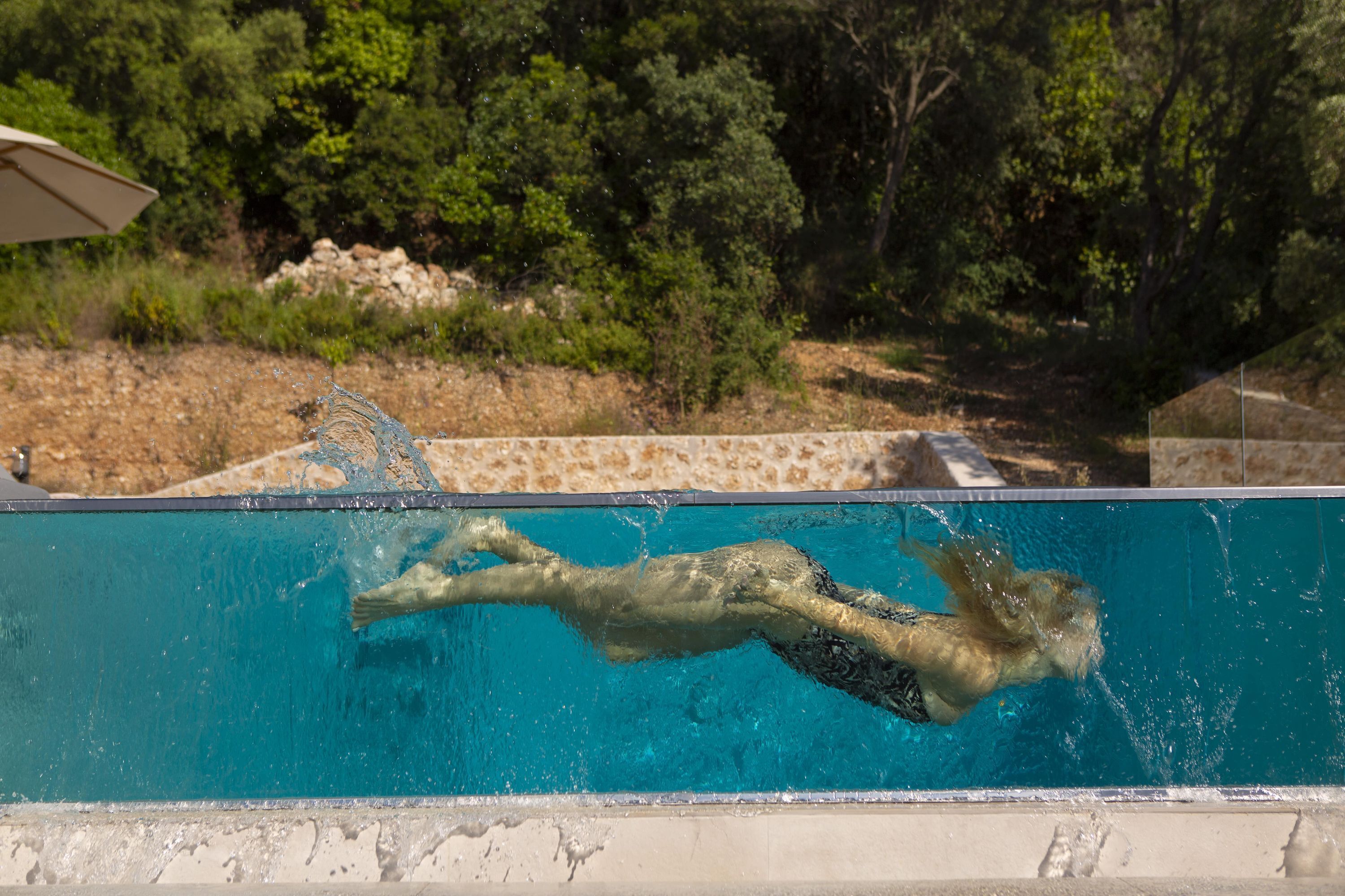 A swimming pool with clear blue water, surrounded by a natural landscape with trees and a stone wall.