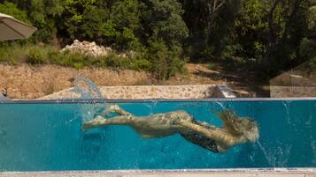 A swimming pool with clear blue water, surrounded by a natural landscape with trees and a stone wall.