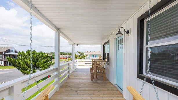 A covered porch with a wooden bench, a table with chairs, and a view of the street and houses. - Newly Updated Cottage in the Heart of KDH! (Kill Devil Hills)
