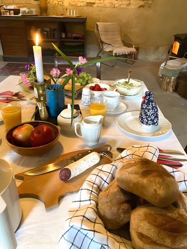 A breakfast table with a candle, fruit bowl, bread, and a vase with flowers.