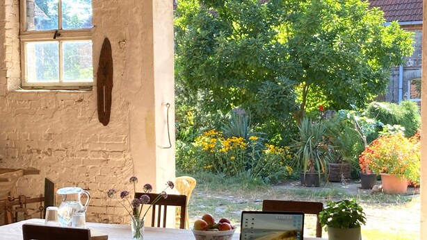A rustic dining area with a table, chairs, a laptop, a bowl of fruit, and a vase with flowers.