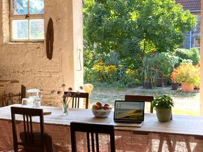 A rustic dining area with a table, chairs, a laptop, a bowl of fruit, and a vase with flowers.