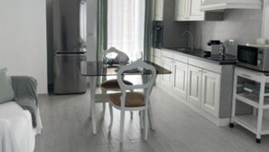 Une cuisine moderne avec des meubles blancs, un réfrigérateur en acier inoxydable, un four à micro-ondes et un coin repas comprenant une table en verre et des chaises blanches.