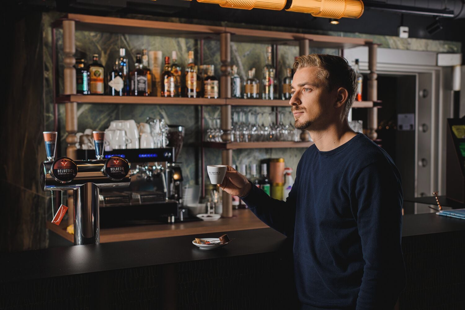 A bar area with a coffee machine, shelves with bottles, and a person holding a cup.