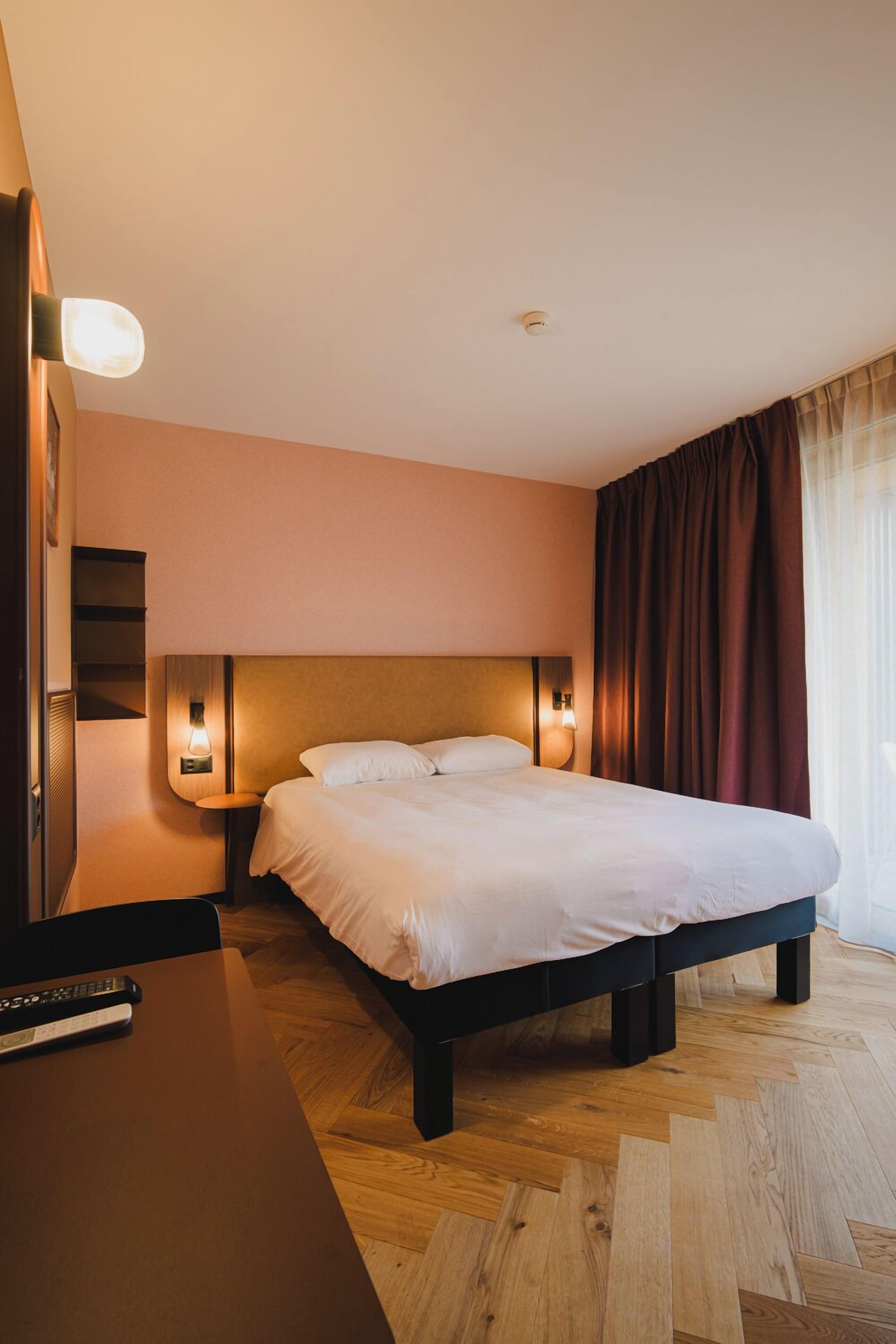 Habitación de hotel con una cama grande, lámparas de noche, un escritorio con un control remoto y una ventana con cortinas.