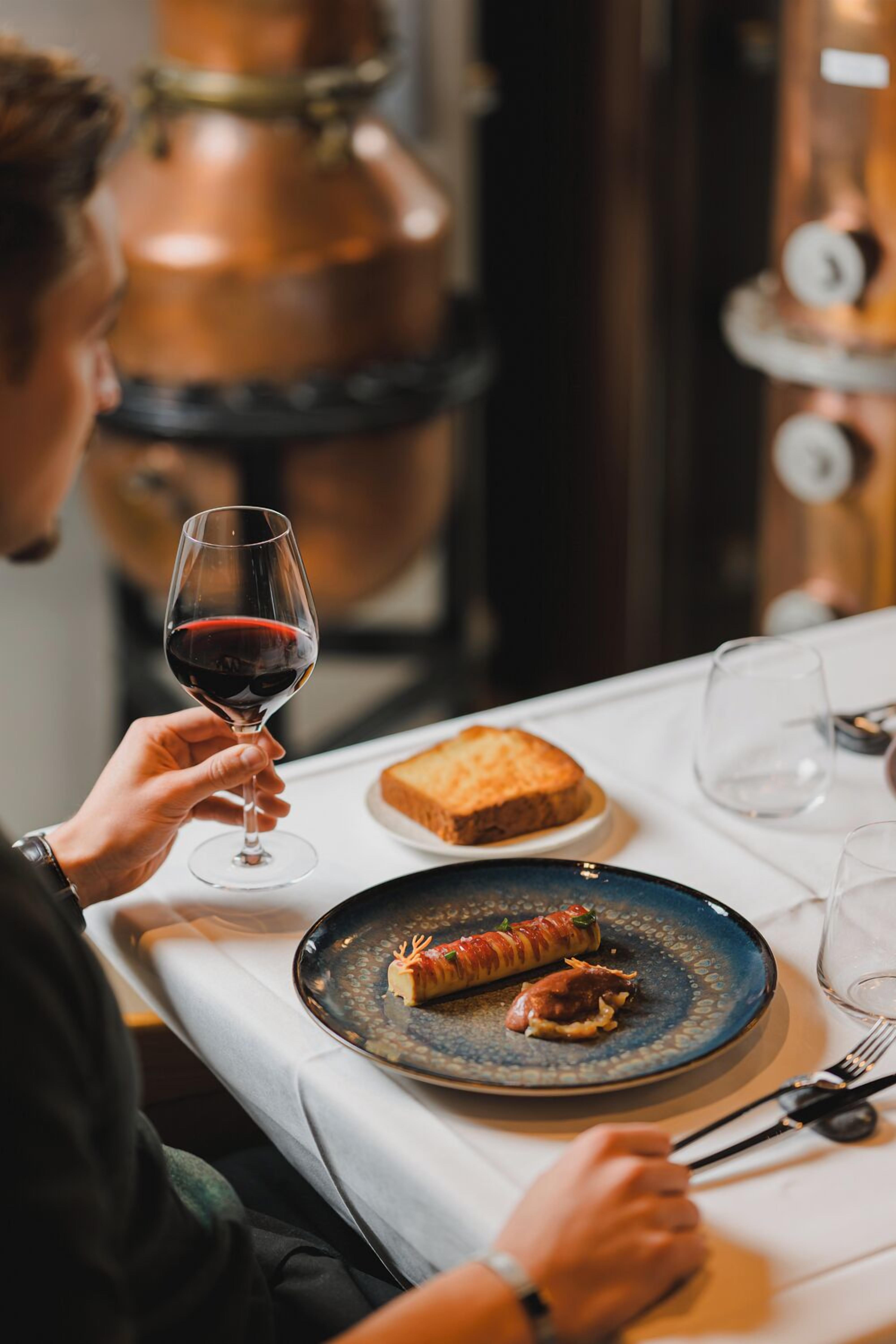 A person holding a wine glass, a plate with food, and a wine bottle in the background.