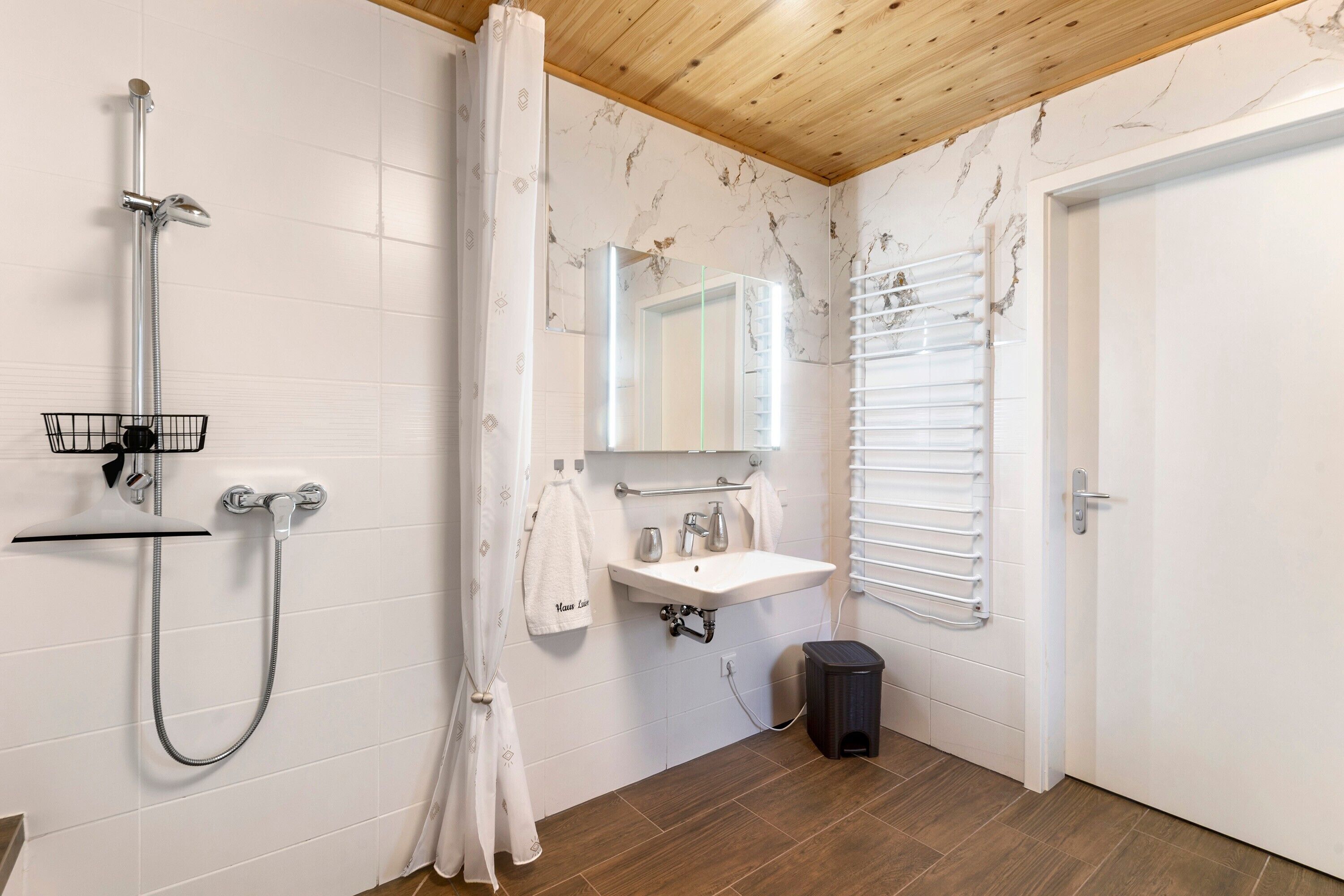 A bathroom with a shower, sink, mirror, towel rack, and a door.