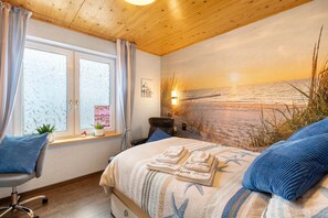 Une chambre à coucher comprenant un lit, une chaise, une fenêtre avec des rideaux, une lampe fixée au mur et une fresque murale représentant une plage.