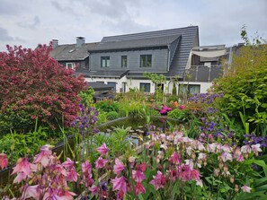 Un immeuble résidentiel doté d’un toit en pente, d’un jardin avec un bassin et de diverses plantes à fleurs.