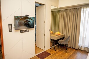 Una habitación con piso de madera, una mesa de comedor con sillas, un televisor montado en la pared y una puerta que da a otra habitación.