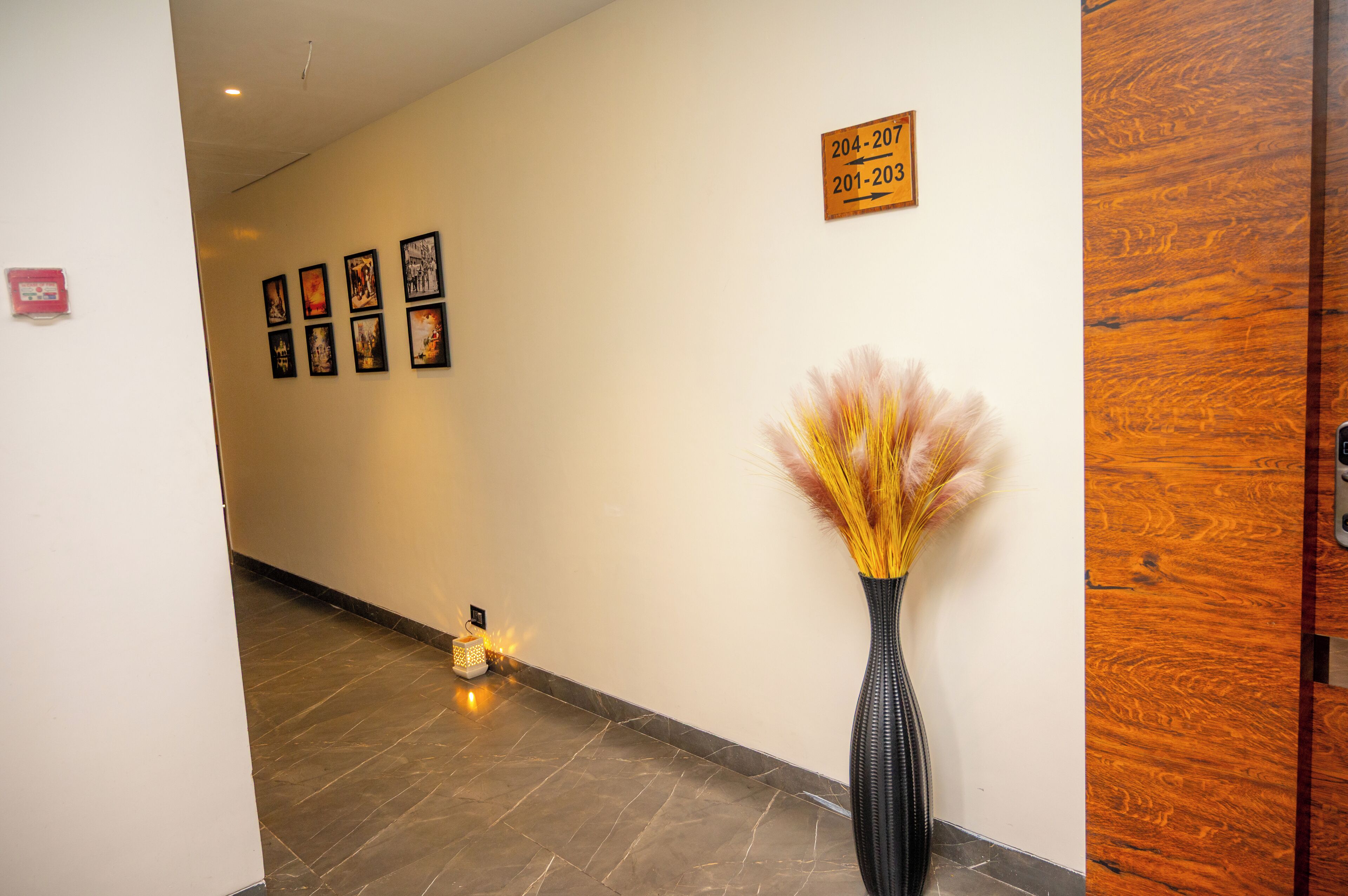 A hallway with a wooden door, a wall-mounted sign with room numbers, and a series of framed pictures.