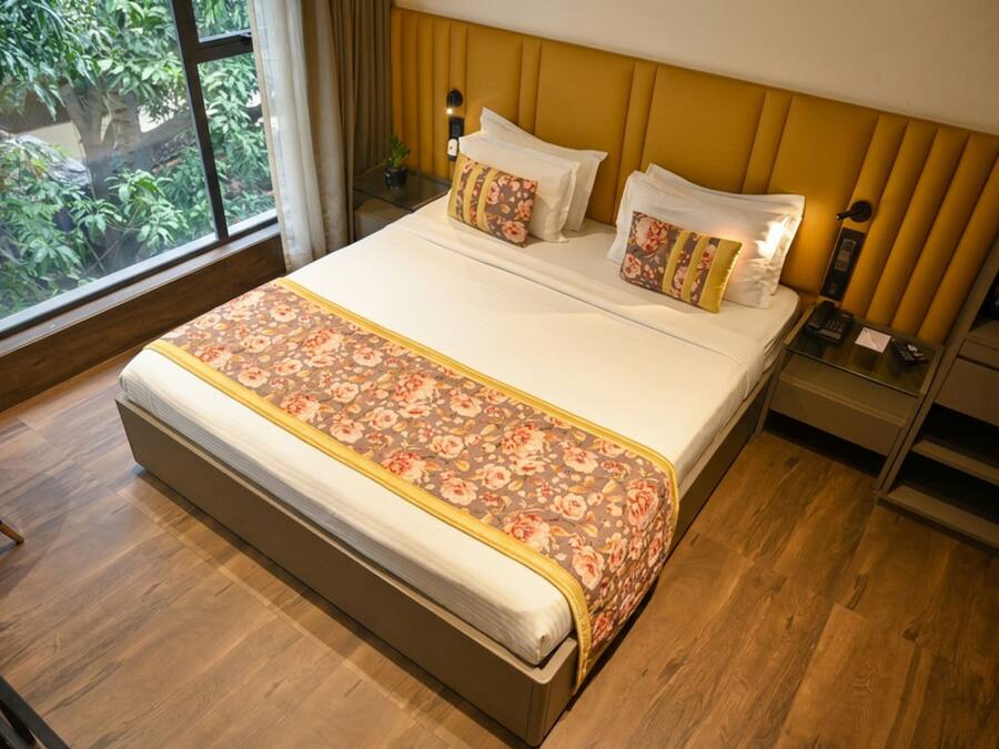 A hotel room with a large bed, wooden flooring, a view of greenery through the window, and a bedside table with a lamp.