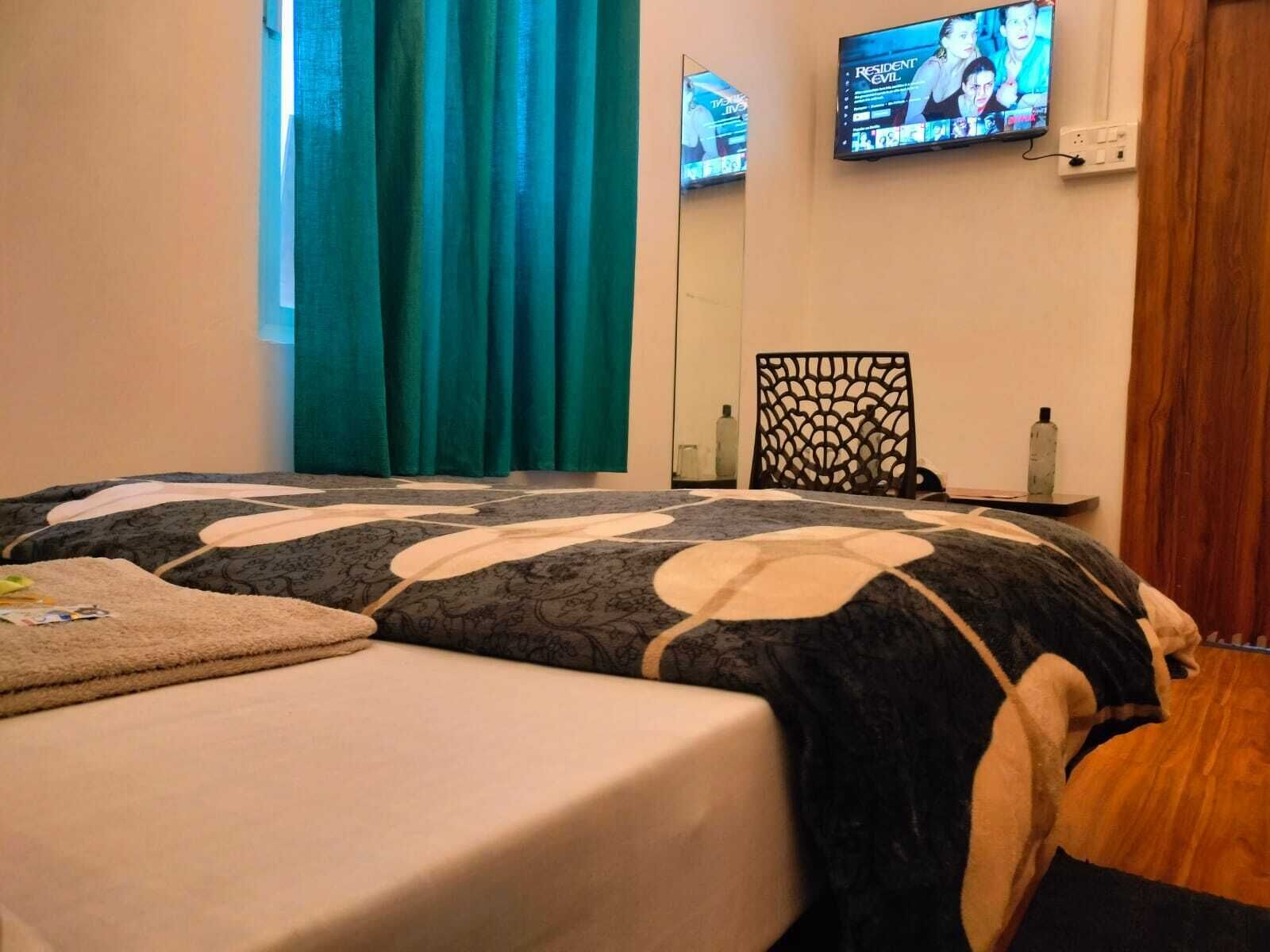 A bedroom with a bed, a TV mounted on the wall, a window with curtains, and a wooden floor.