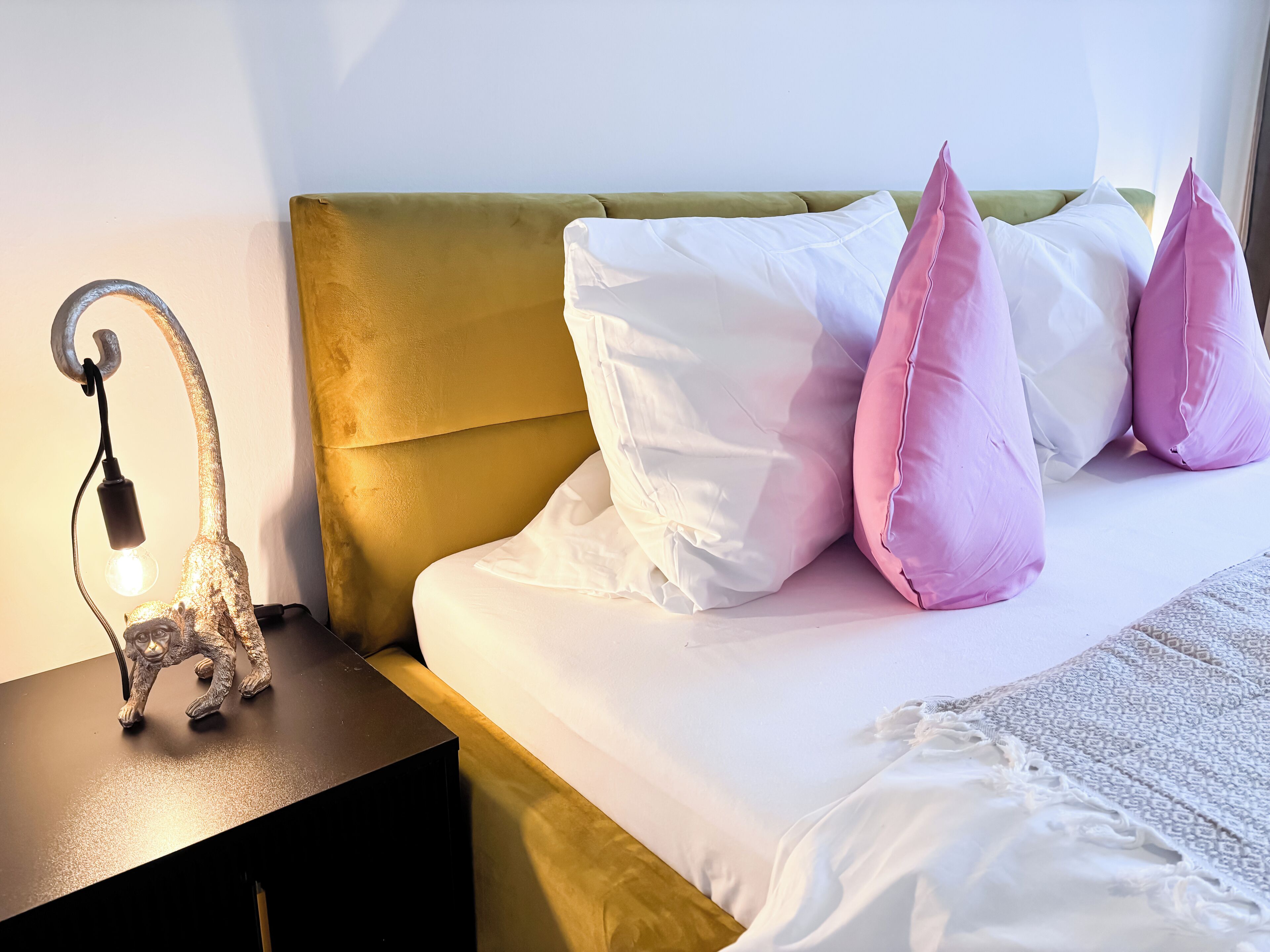 A neatly made bed with white and pink pillows, a gold headboard, and a small table with a decorative lamp.