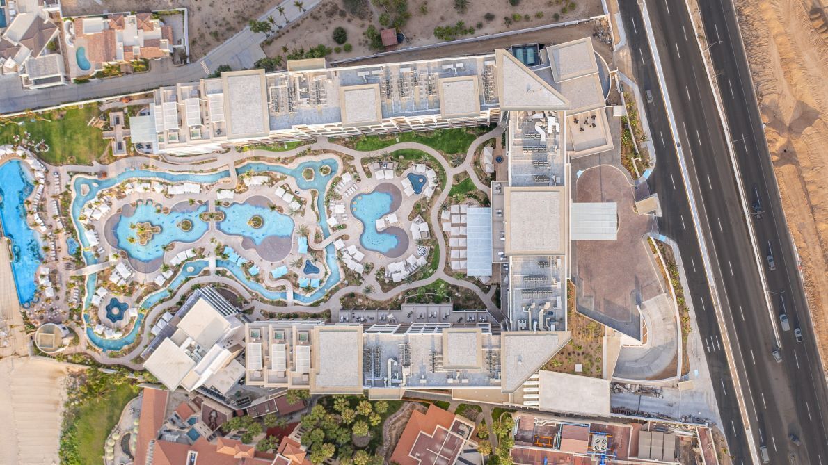 Una vista aerea di un hotel con diverse piscine, un ampio parcheggio e una strada adiacente alla struttura.