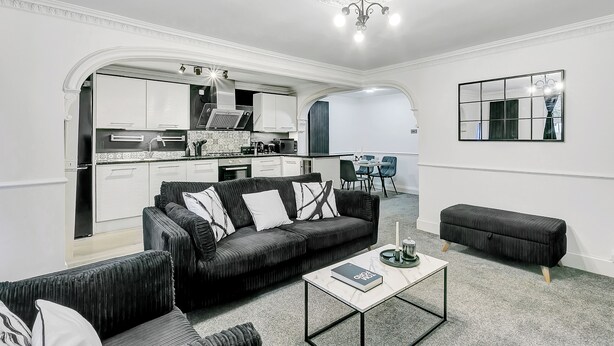 A modern living room with a black sofa, a coffee table, and a dining area in the background.