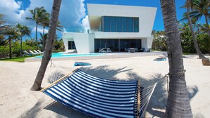 A modern building with a pool, surrounded by palm trees and a hammock. - Olympus: Luxe Villa w/ Designer Pool, Private Beach & Rooftop Lounge w/ Panoramic Views (Grand Cayman)