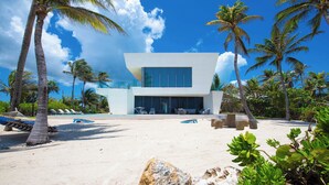 A modern building with a large glass facade, surrounded by palm trees and a sandy area with lounge chairs. - Olympus: Luxe Villa w/ Designer Pool, Private Beach & Rooftop Lounge w/ Panoramic Views (Grand Cayman)