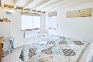 Une chambre à coucher avec un lit, deux fenêtres avec des rideaux, un radiateur, une chaise et une étagère avec des objets.