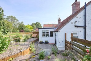 A white house with a red-tiled roof, a wooden fence, and a garden with various plants and a bench. - Host Stay Pebble Cottage (Norwich)