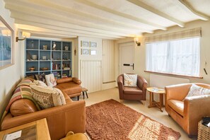 A cozy living room with a brown leather sofa, a small armchair, and a coffee table. There is a bookshelf filled with books and decorative items, and a window with sheer curtains. - Host Stay Pebble Cottage (Norwich)