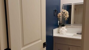 A bathroom with a white door, a blue accent wall, a mirror, a sink, and a vase with flowers. - Cozy Haven in Glenbrook close to downtown (Calgary)