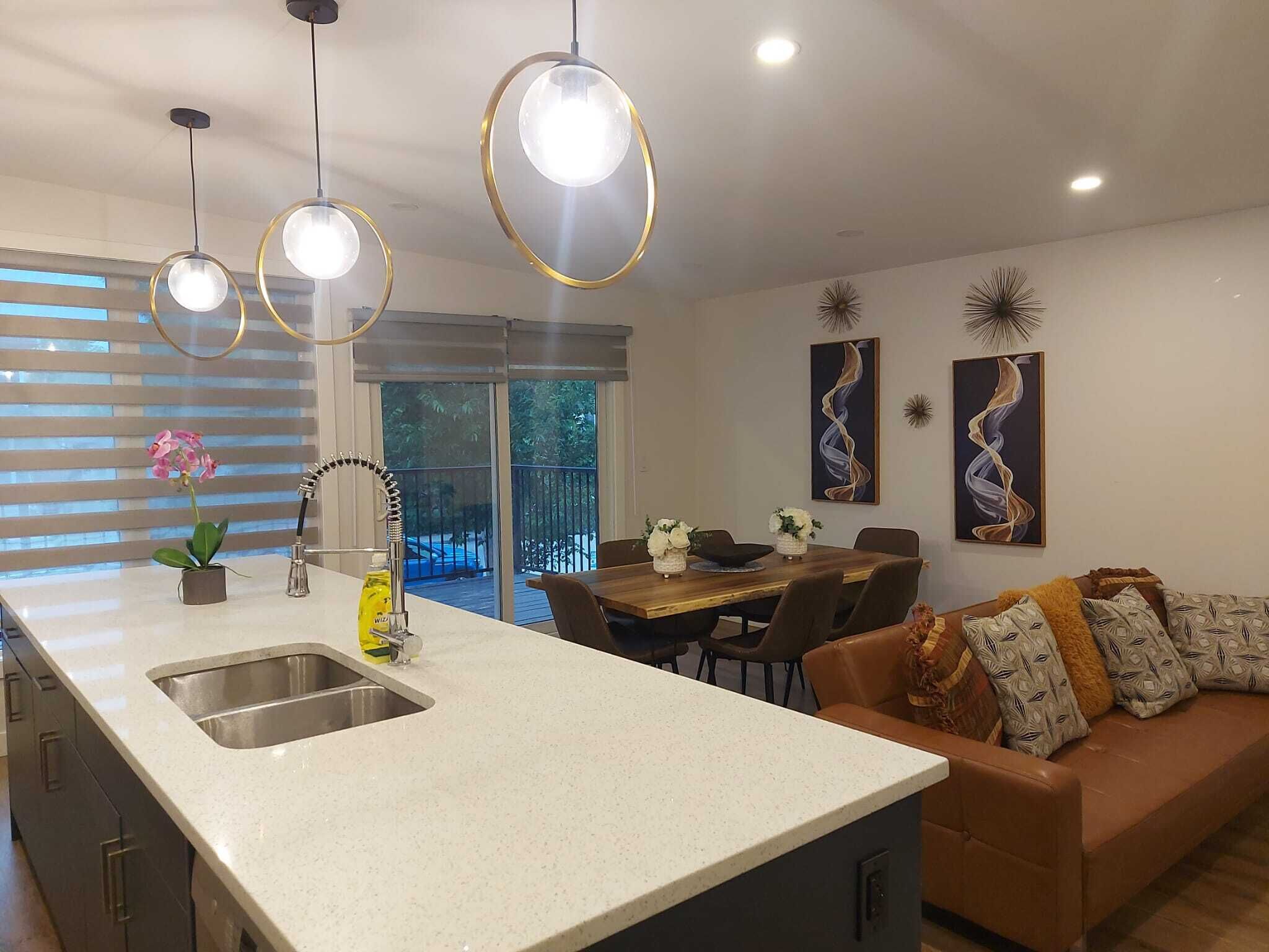 A modern kitchen with a large island, a dining area with a wooden table and chairs, and a living area with a sofa and cushions.