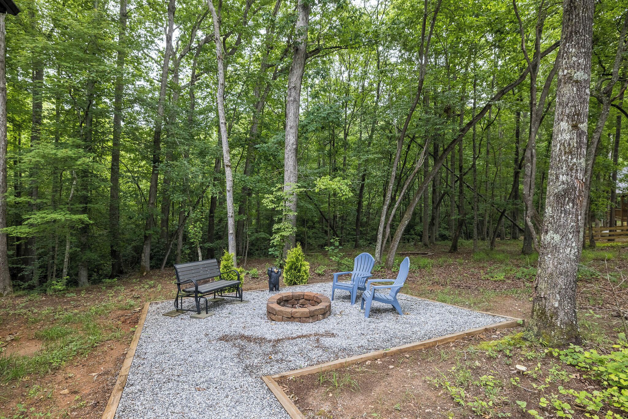 A forested area with a fire pit, two Adirondack chairs, and a bench.