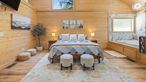 A wooden cabin bedroom with a bed, two bedside lamps, a window, a painting, a potted plant, and a bathroom with a bathtub and sink. - Prized Gem - Spa-like Retreat in the Heart of Nature (Cleveland)