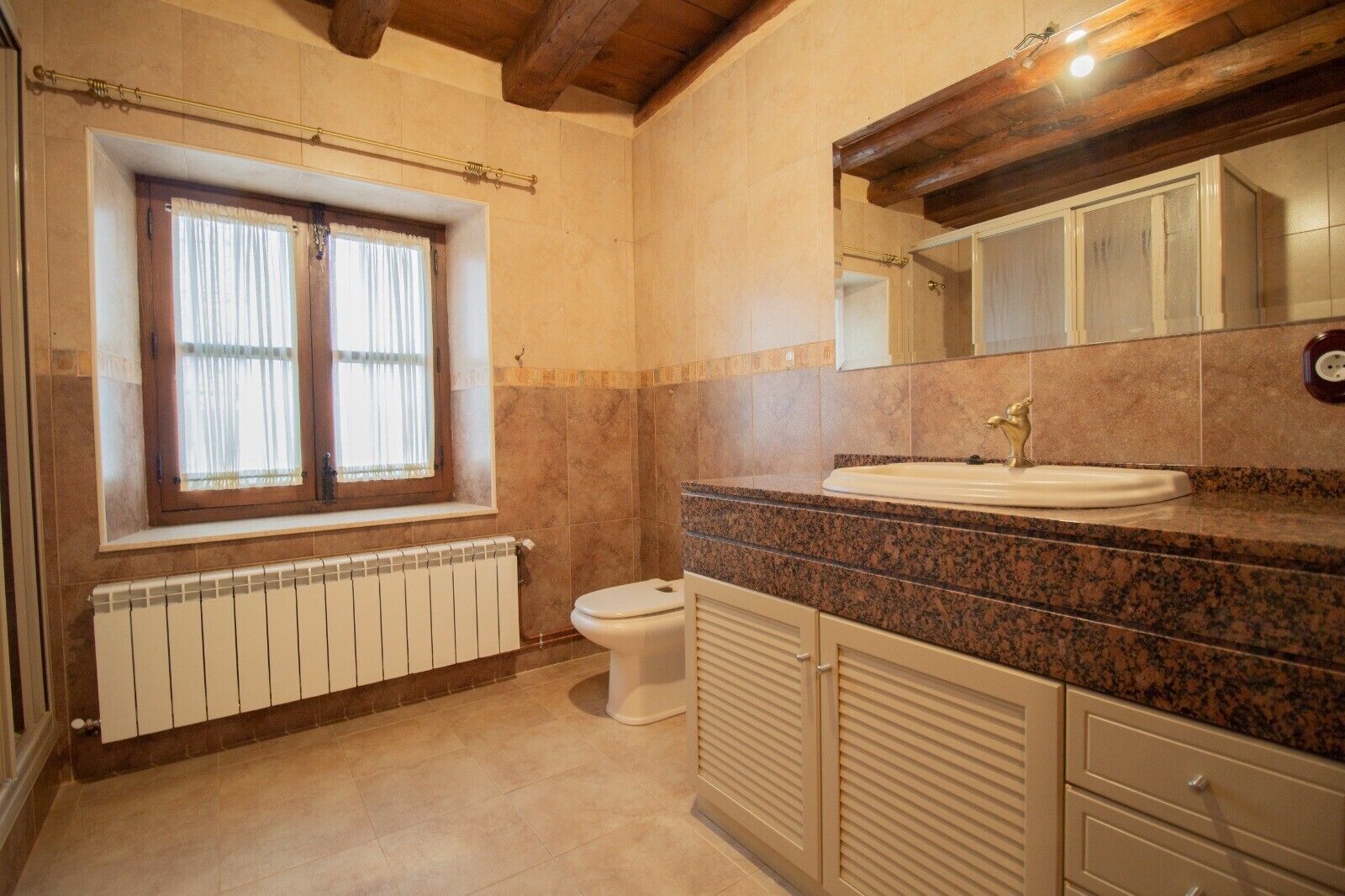 Un baño con ventana, radiador, inodoro y lavabo con espejo superior.