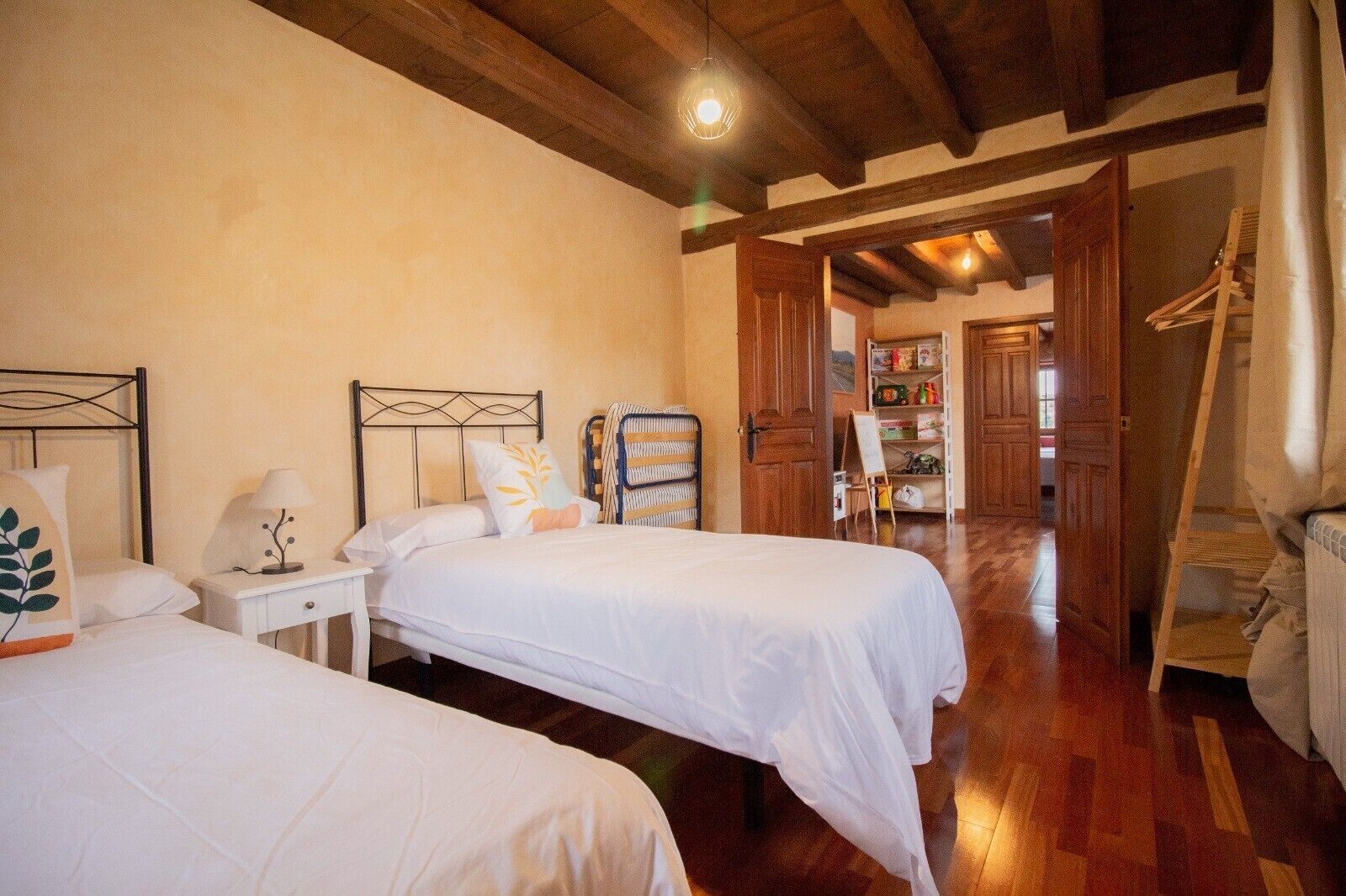 A room with two beds, wooden flooring, a wooden ceiling, and a view of a hallway with a door leading outside.