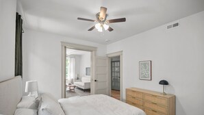 A bedroom with a bed, bedside table, lamp, dresser, ceiling fan, and a door leading to another room.