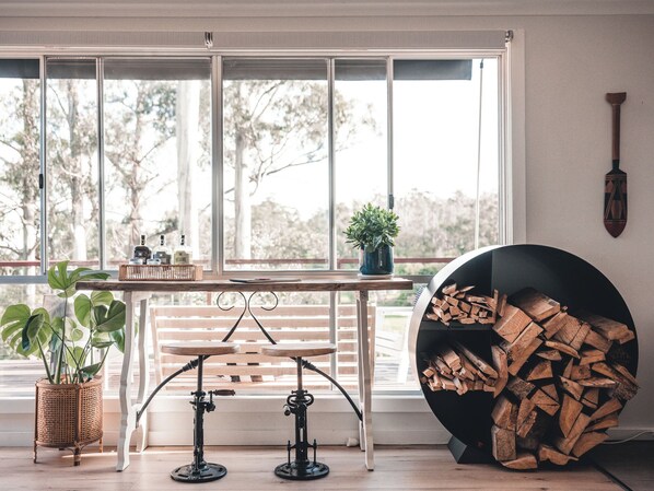 A room with a large window, a table with bottles, a potted plant, a metal stool, and a wood-burning stove.