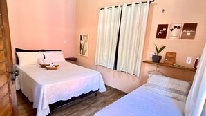 Habitación de hotel con dos camas, una ventana con cortinas, un equipo de aire acondicionado y un estante pequeño con una planta y marcos de fotos.