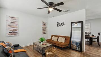 Una sala de estar moderna con un sofá marrón, un sofá negro, una mesa de centro de madera y un ventilador de techo. Se puede ver un comedor a través de una puerta de cristal.