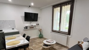 A compact room with a bed, a dining table with chairs, a small round table, a TV mounted on the wall, and a window with curtains. - T2 apartment for rent (Chorges)