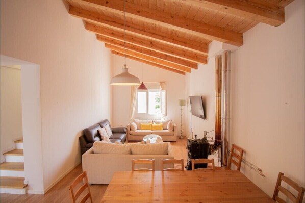 A living room with a wooden ceiling, a sofa set, a dining table with chairs, and a TV mounted on the wall.
