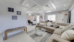 A modern living room with a beige sofa, a glass coffee table, and a patterned rug. There is a bench against the wall and a dining area in the background. - 3 Mi to Beach: Home w/ Outdoor Space in Hollywood (Hollywood)