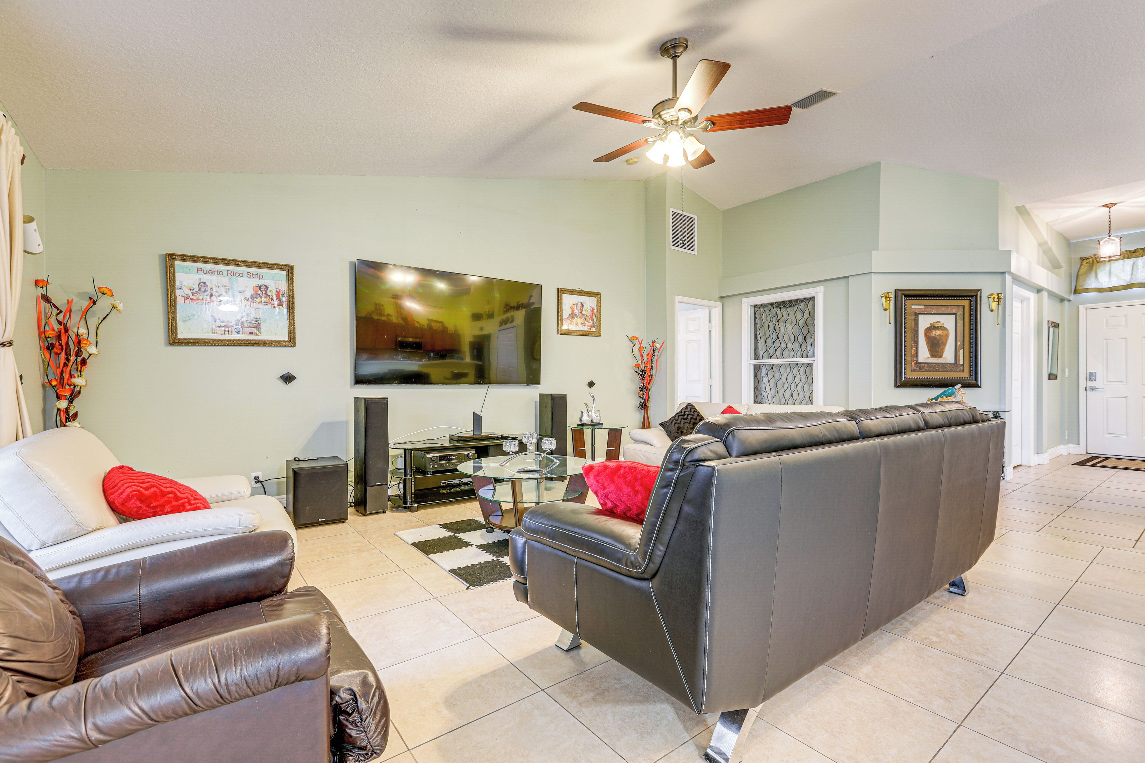 A living room with a sofa, a coffee table, and a TV. There is a ceiling fan, a framed picture on the wall, and a door in the background.