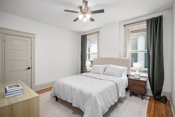 Un dormitorio con cama, mesitas de noche, ventilador de techo y ventana con cortinas.